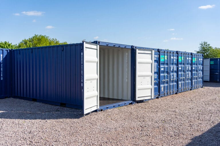 Blue self storage containers at Optima Self Store Newark, with one unit open to show the empty storage space and gravel access area.