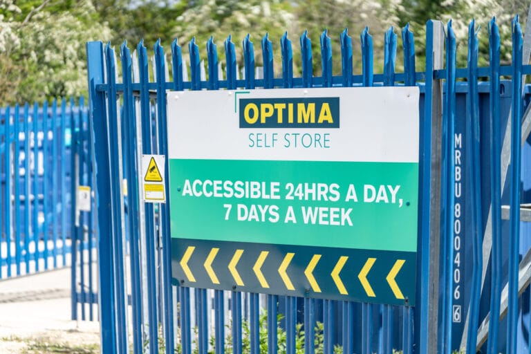 Optima Self Store sign on a blue security fence advertising 24 hour access, seven days a week, at the Stamford storage facility.