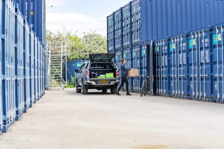 Person carrying a cardboard box beside a parked pickup truck between rows of blue storage containers at Optima Self Store Stamford.